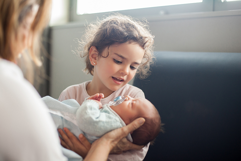 A little girl holding a small baby. She looks down at the baby's face. The mothers hand is supporting the baby's head. The baby sucks a blue and white soother.