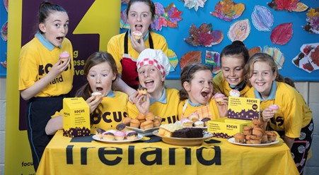 School bake sale in aid of Focus Ireland. There is a group of kids wearing Focus Ireland t-shirts behind a table. There are lots of cakes on the table.