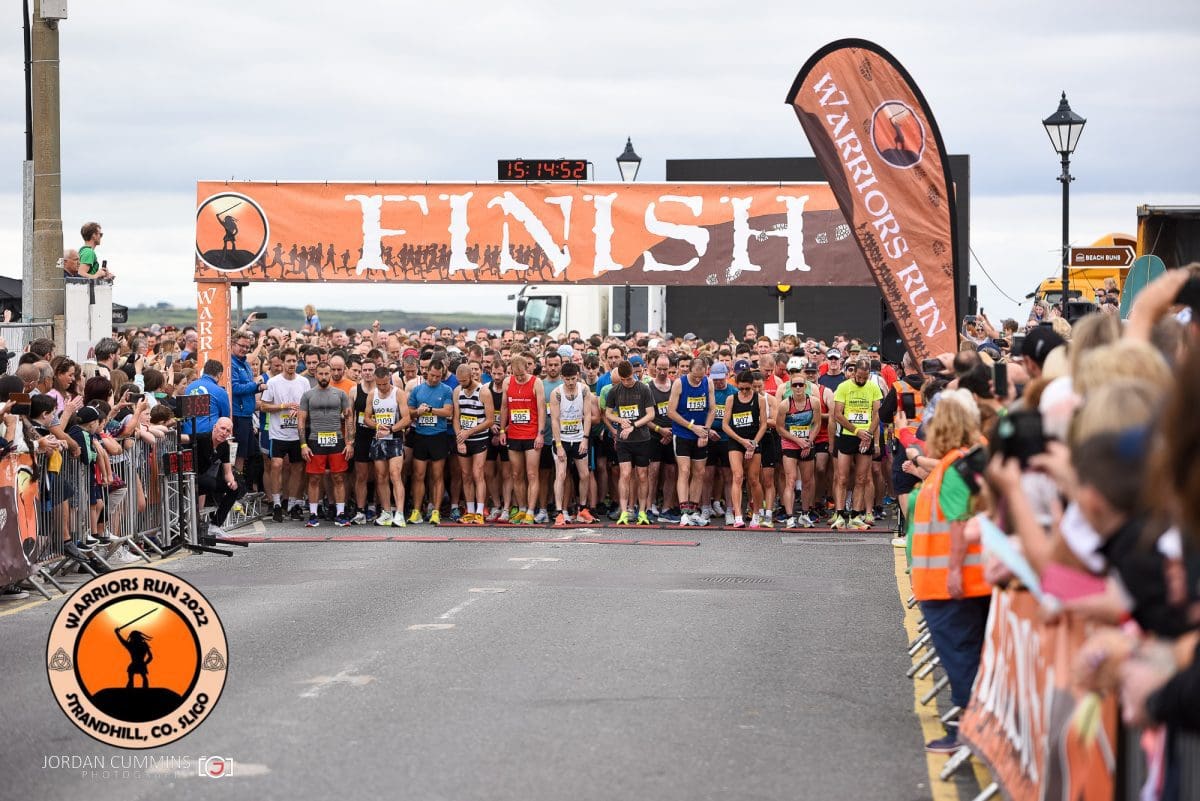 Participants in the Warriors Run 2022 are lined up at the at the start line. They all wear running gear. There are spectators behind silver barriers.