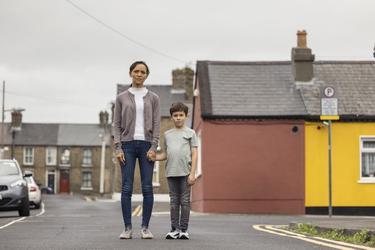 A mother and child stand together holding hands outside. There are houses and cars in the background.