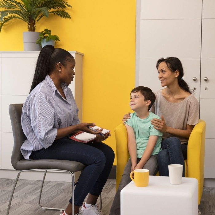 A mother and her son speak to a member of Focus Ireland support staff. They sit on the same yellow chair. The mother has her hands resting on her sons shoulders.