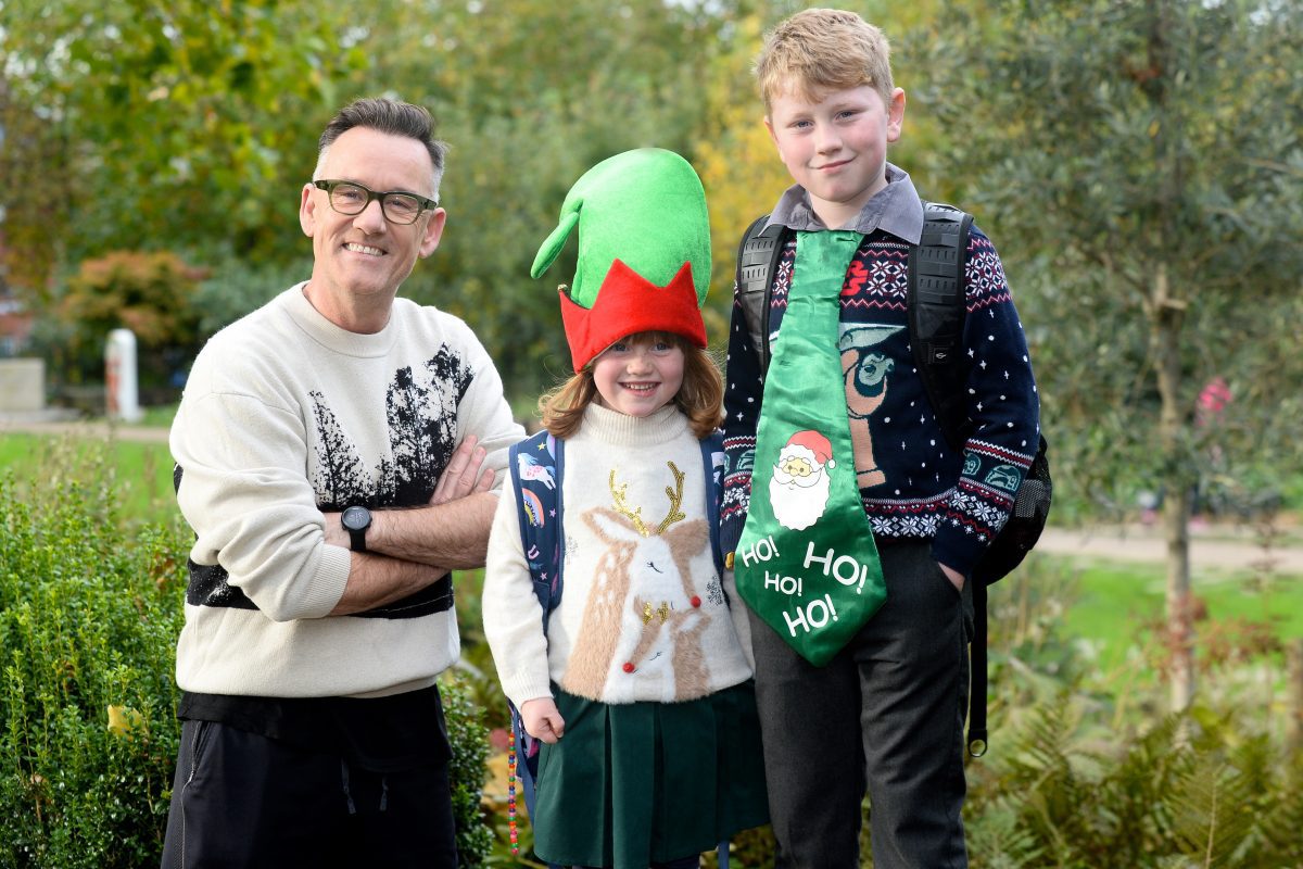 Brendan Courtney smiles to camera next to two children. They are all wearing Christmas jumpers and smiling to camera.