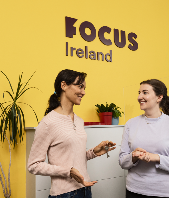 Two woman smiling at each other. There is a yellow wall with the purple Focus Ireland logo on it.
