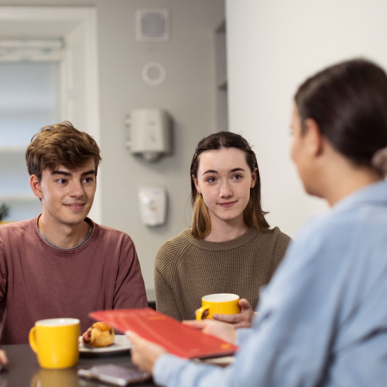 An over the shoulder view of a Focus Ireland key worker having coffee with man and a women. The key worker holds a red copy.