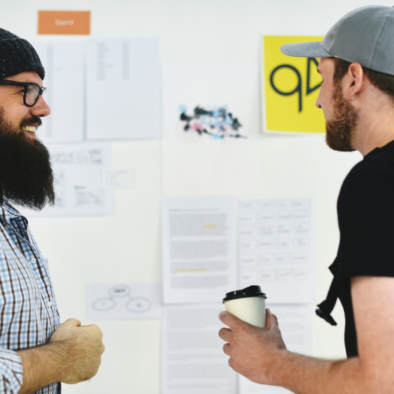 There are two men infront of posters. A man wearing a cap has his back facing the camera. The other man smiles towards him.