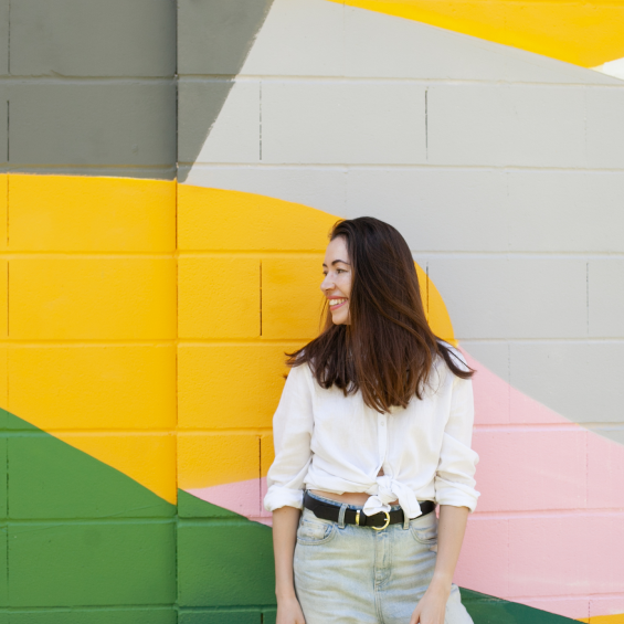 There is a woman standing infront of a yellow, grey, pink, and green stripped wall. She is smiling and looking to the left.