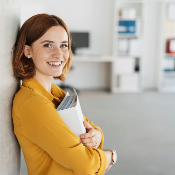 woman smiling to the camera with her back against the wall. She is holding on her chest a file folder.
