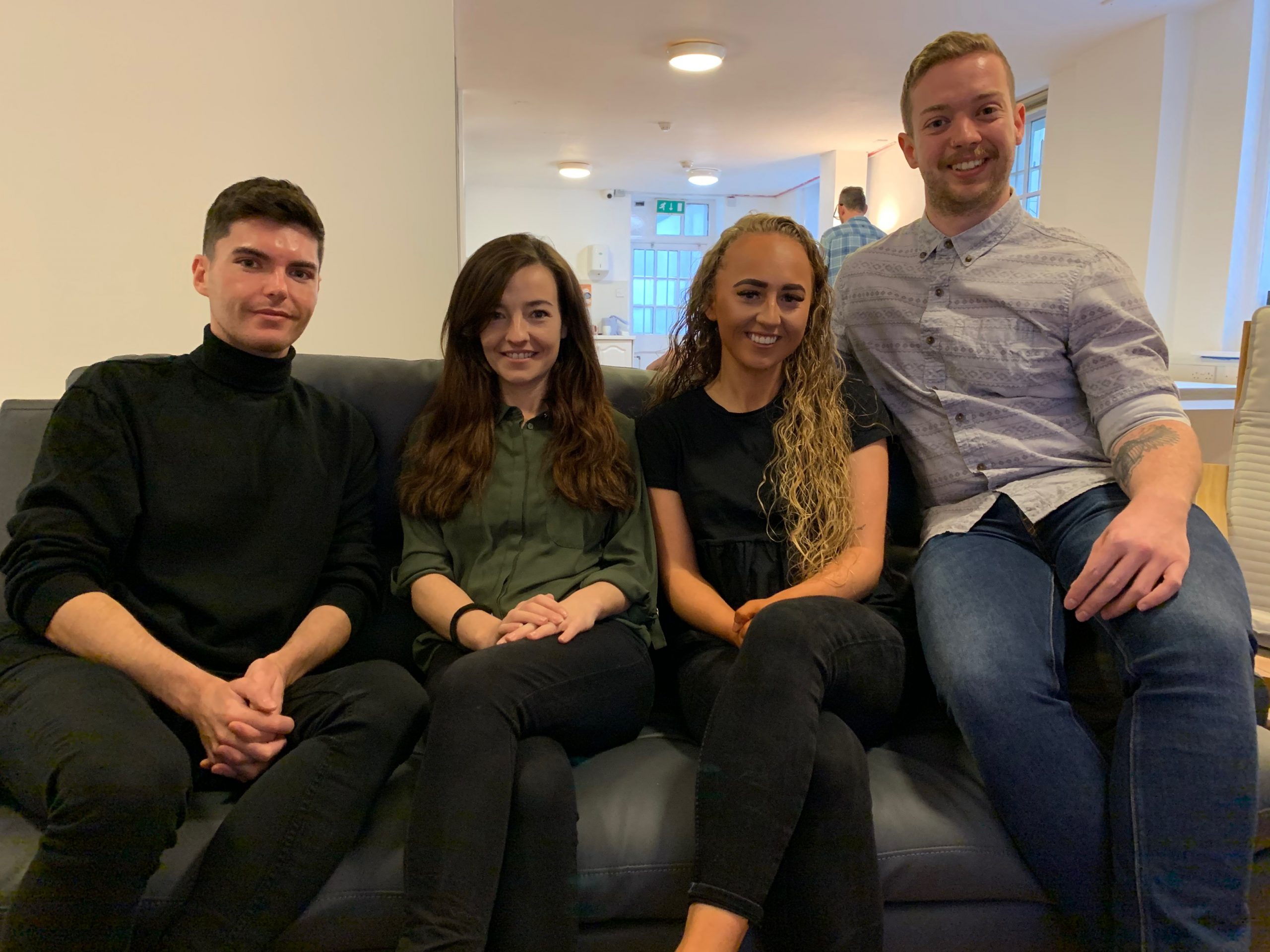 Four people sitting on a black couch. There is a man facing away in the background.