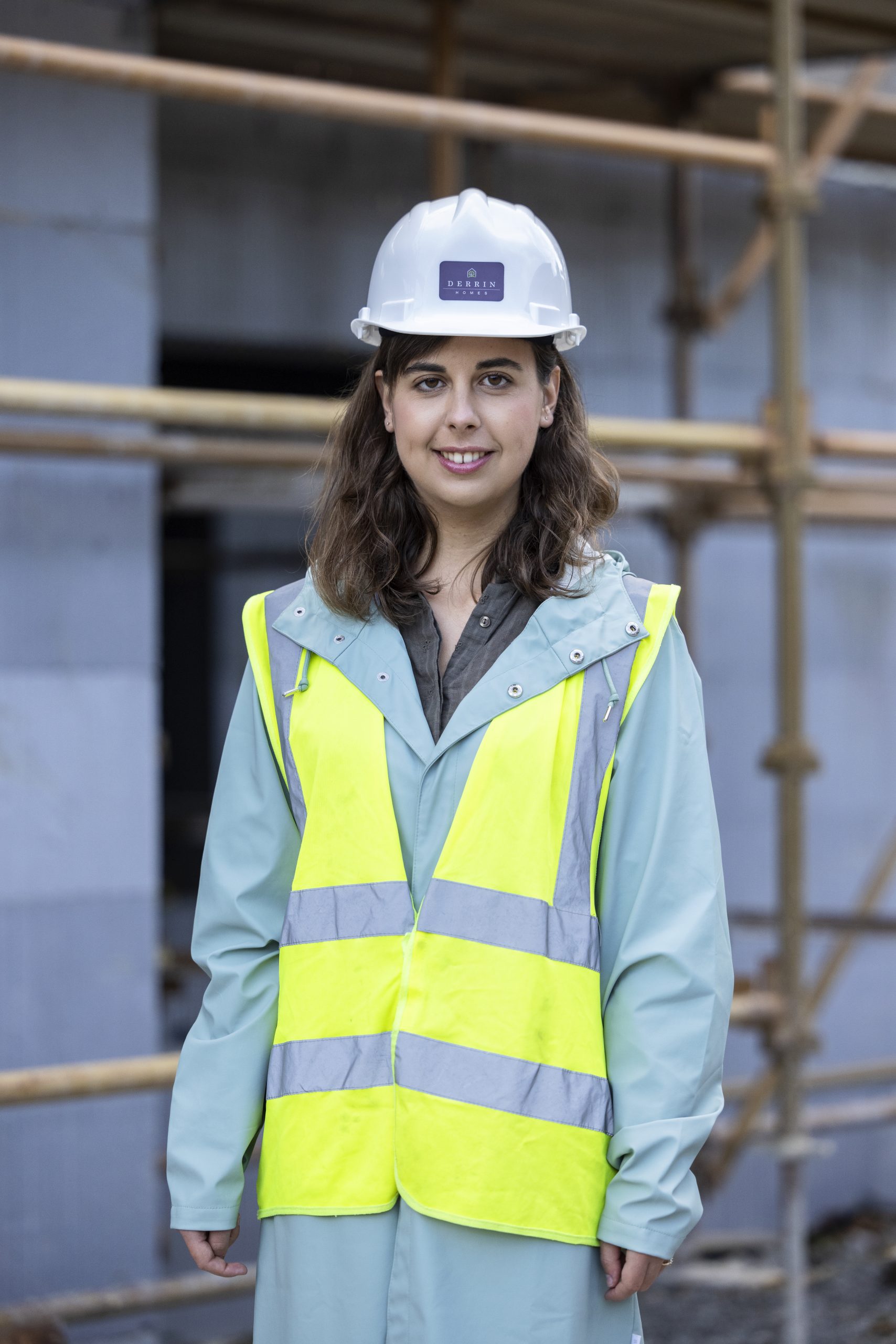 Focus Ireland staff member visits a building site. She is wearing a Hi-vis jacket and a hard protective helmet over her long blue coat. There is a building and scaffolding behind her.