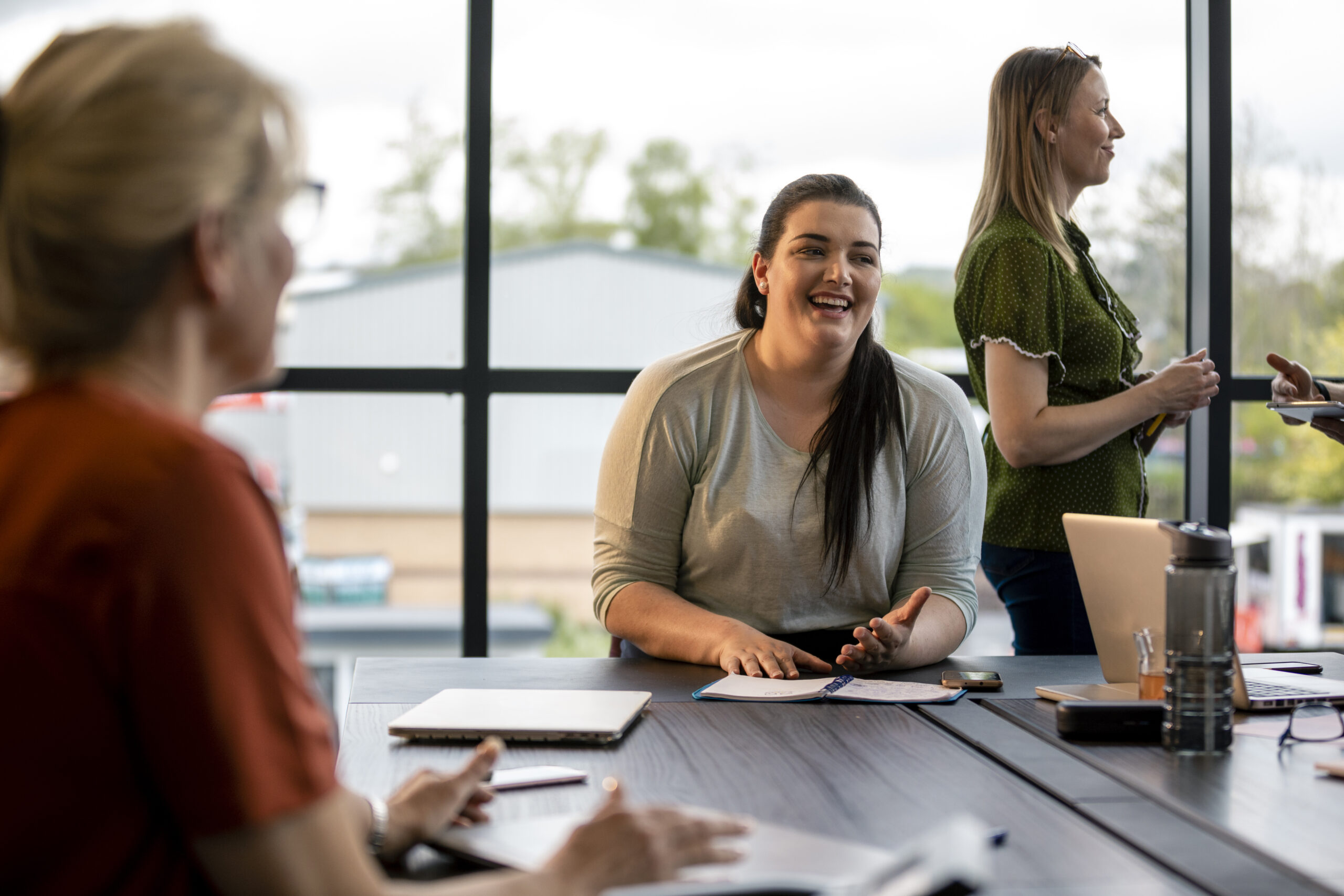 Over the shoulder view of the board room in Hexham. One staff member is laughing as she speaks with her colleagues. Another staff member stands smiling at someone in the background. There is a laptop notebooks and phones on the table.
