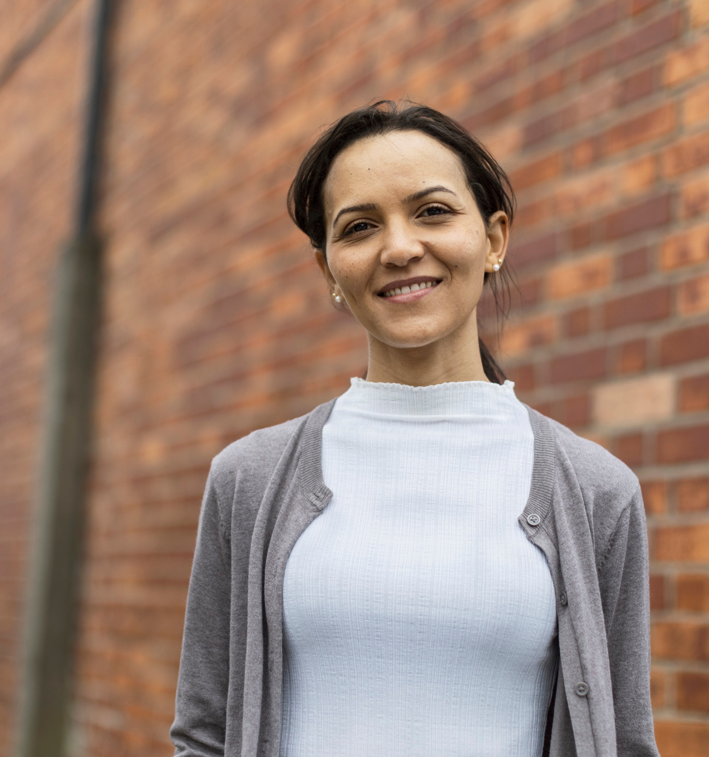 A woman stands smiling towards the camera. There is a red bricked wall with a pipe running down it behind her.