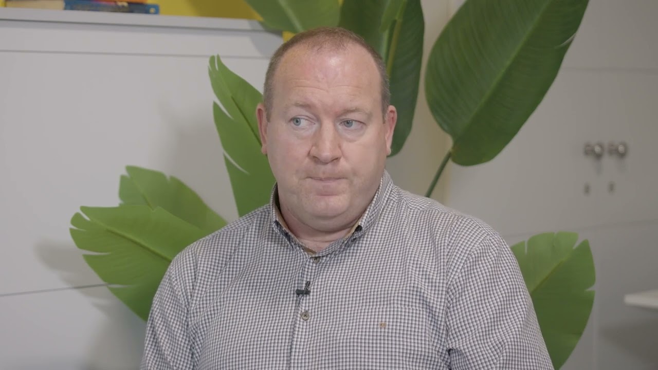 A man is looking to the left as he speaks. He is wearing a checkered shirt. There is a white cabinet and a large plant behind him.