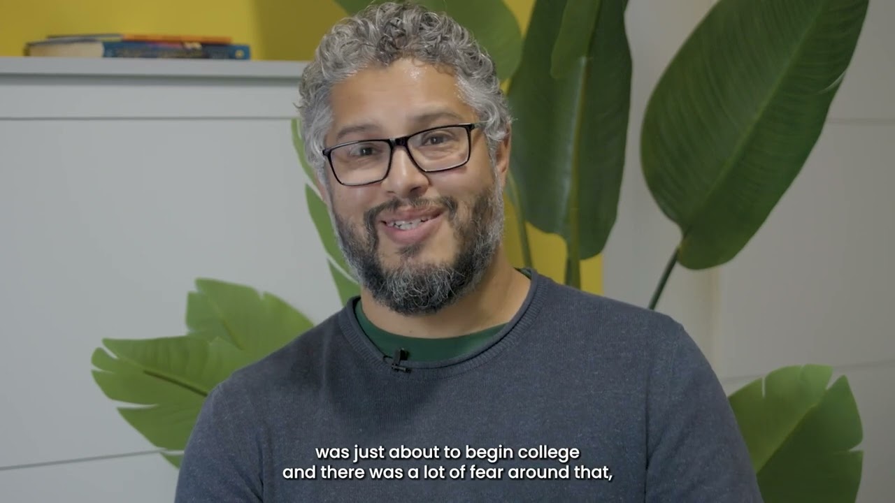 A man with grey curly hair, a dark beard and glasses has a smile on his face. He is speaking to the camera. There is a white cabinet and a plant behind him.
