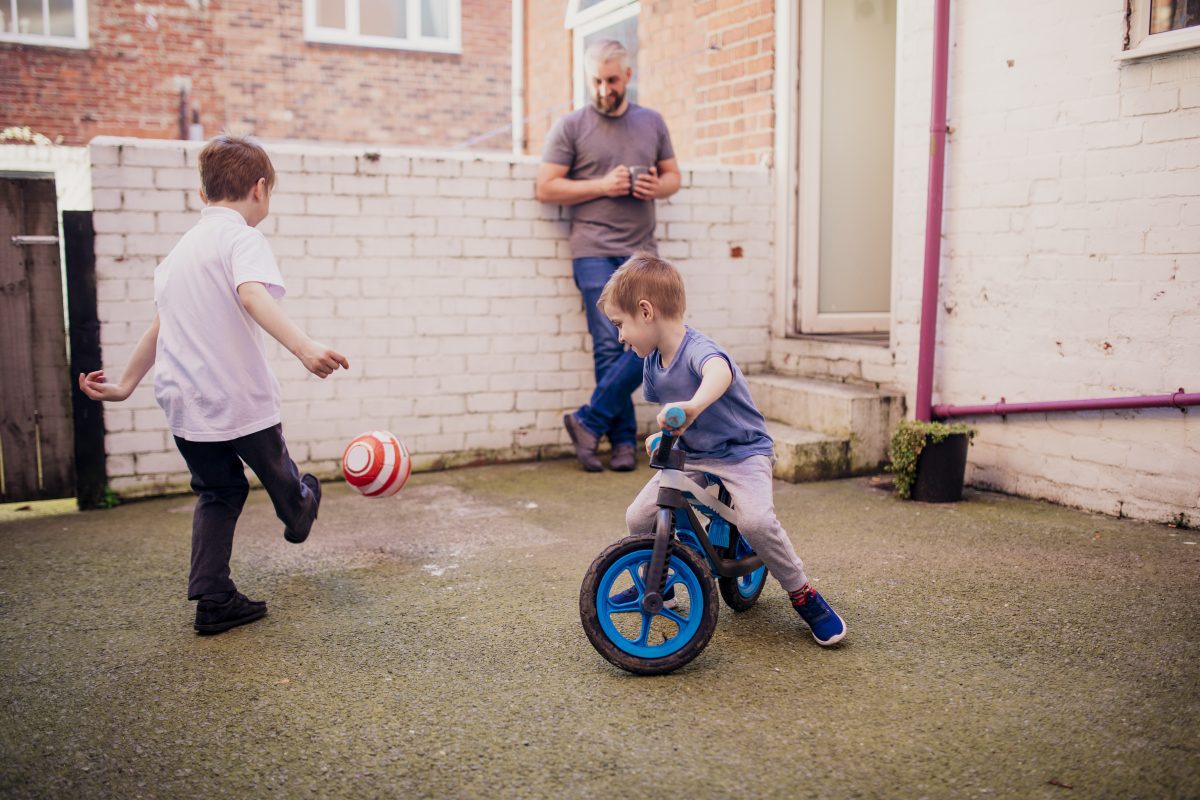 A father watches as his two sons play. One kicks a football towards a brick wall painted white, while the other is riding a bike.