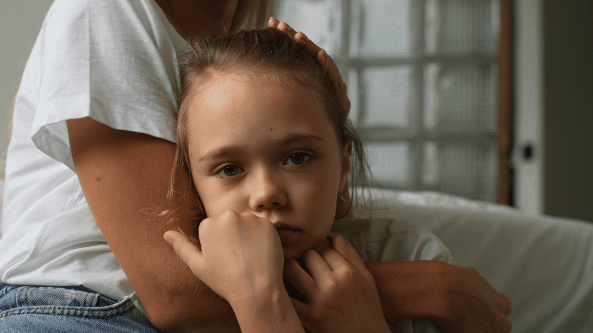 A child looks to camera. They are holding on to their parents arm who is hugging them. The parents face is out of frame.