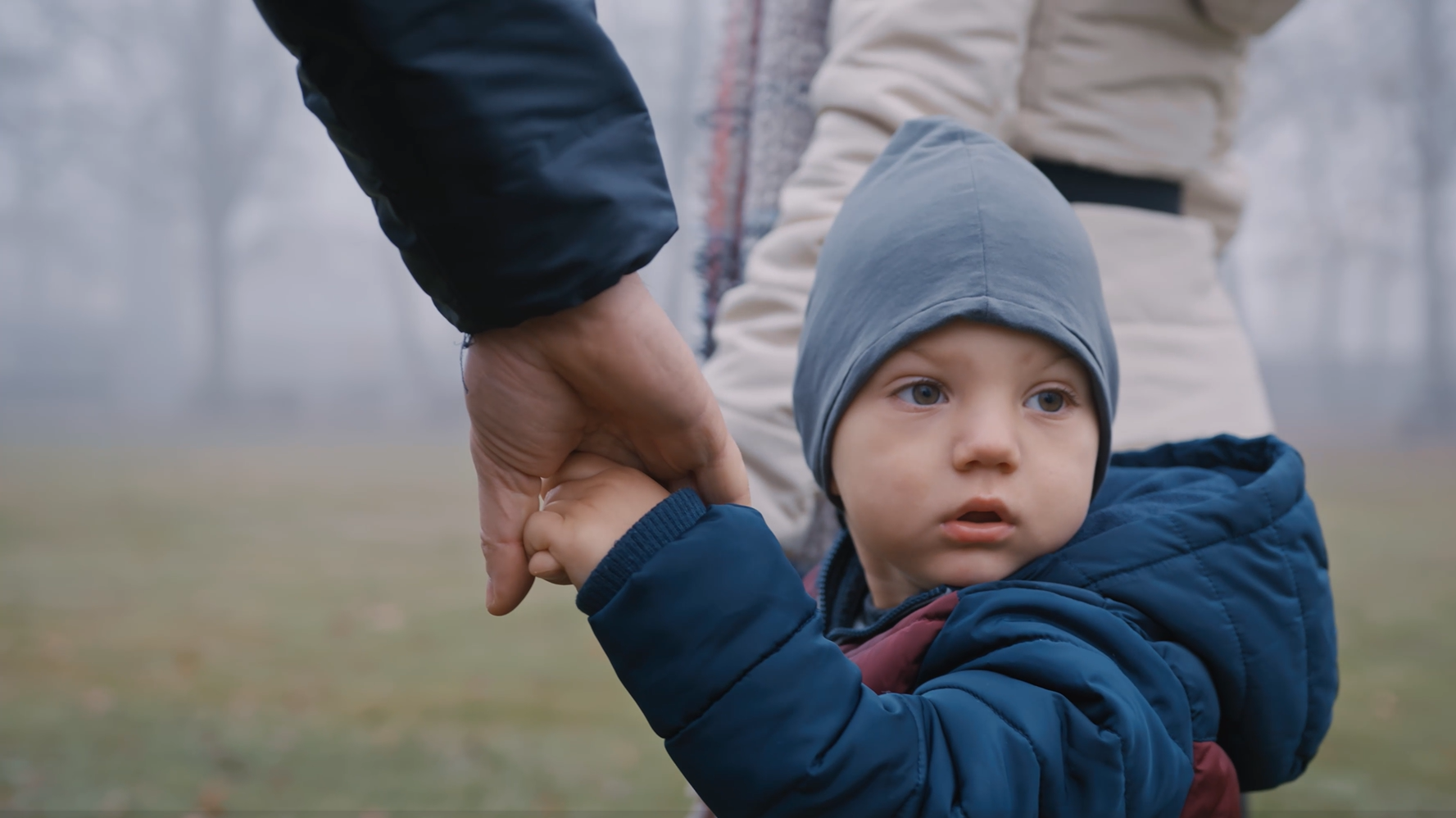 A young child looks over their shoulder as they hold their parents hands. They are are outside on a foggy day. They are wearing a heavy jacket and a woolen hat.