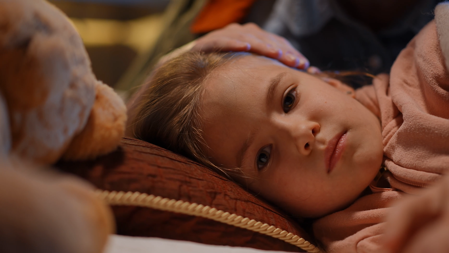 A child lays on her side, with her head resting on a pillow. A parents hand is resting gently on her head.