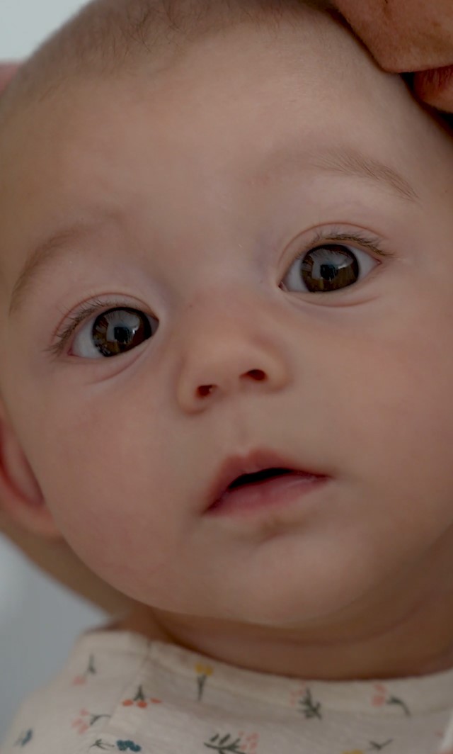 A baby looks at the camera. Their head is being cradled by their mother's hand.