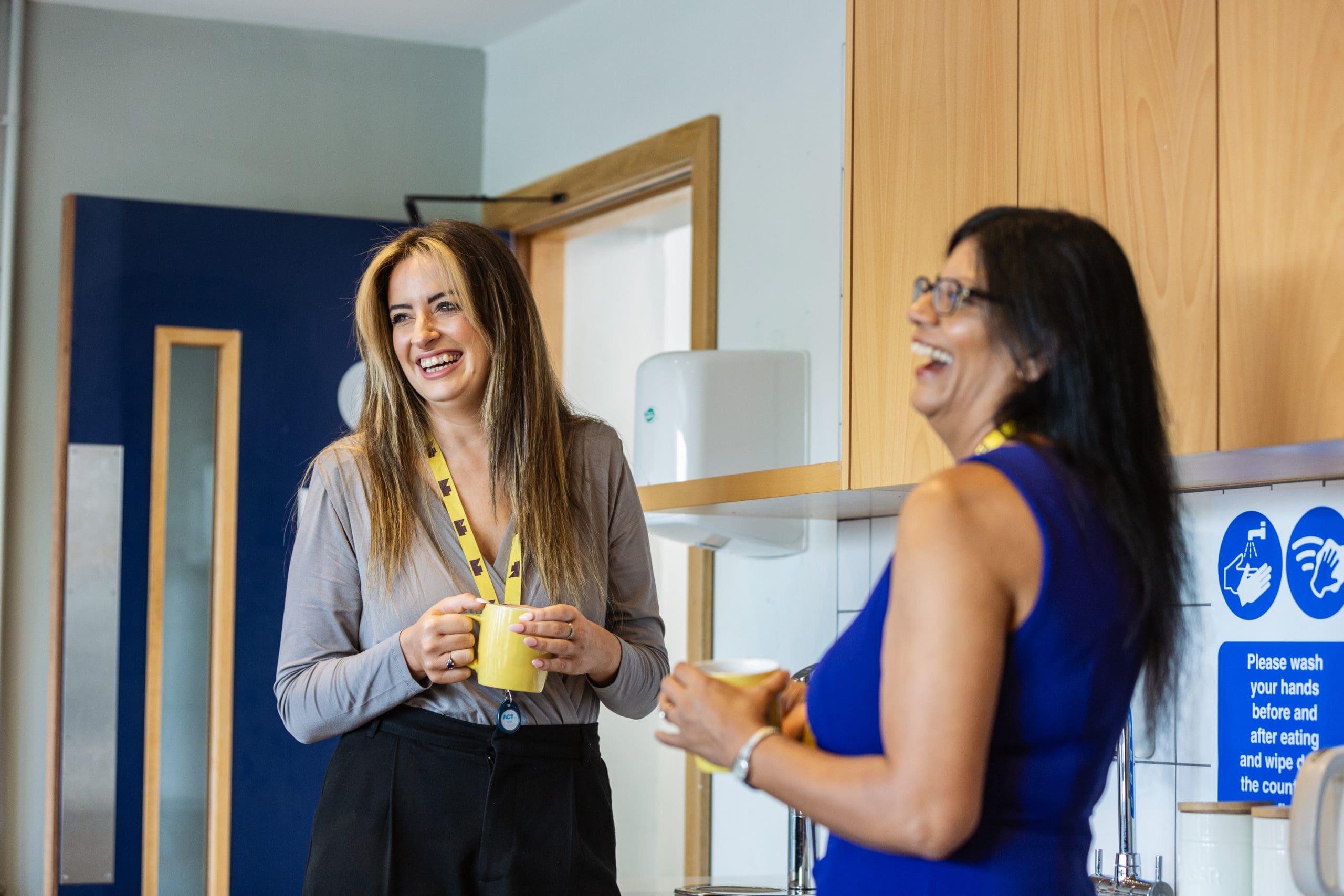 Two Focus Ireland employees stand in an office kitchen. They are looking out of frame and laughing.