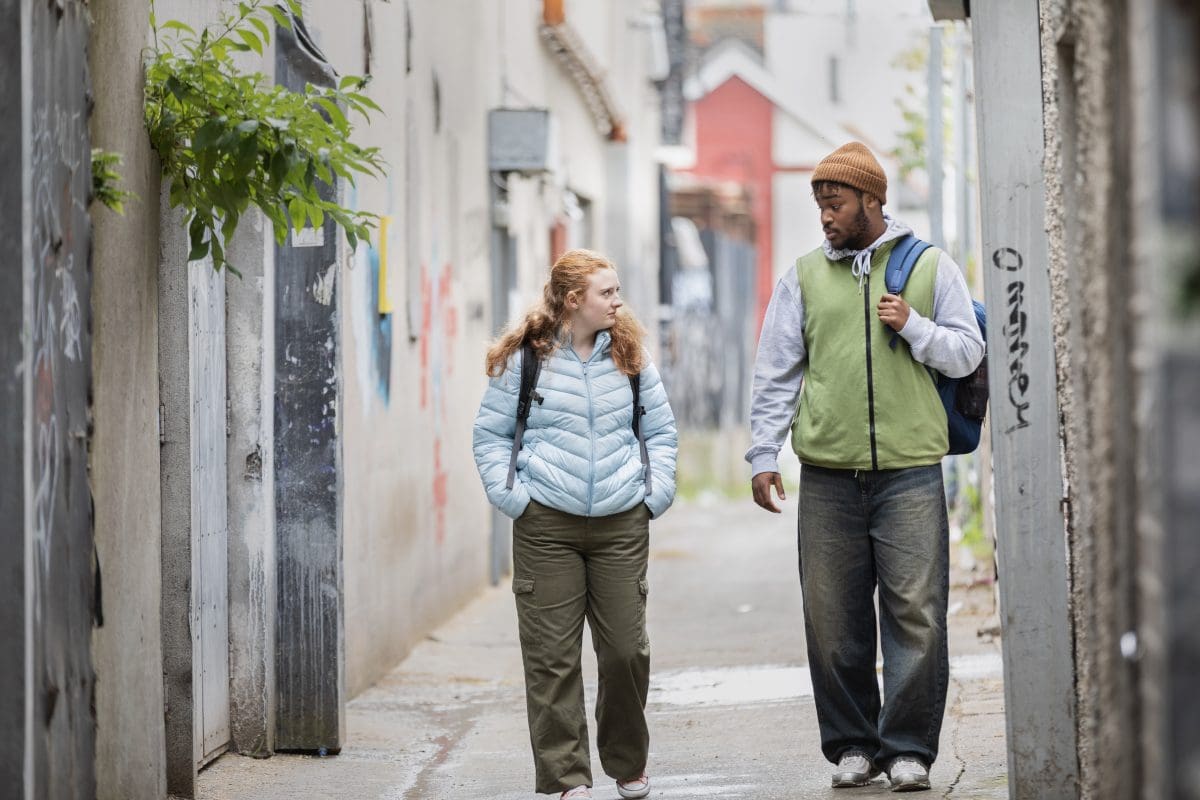 Two young people walk through a ide street with backpacks on their back. They are talking to each other/ Both of them are wearing warm clothes.