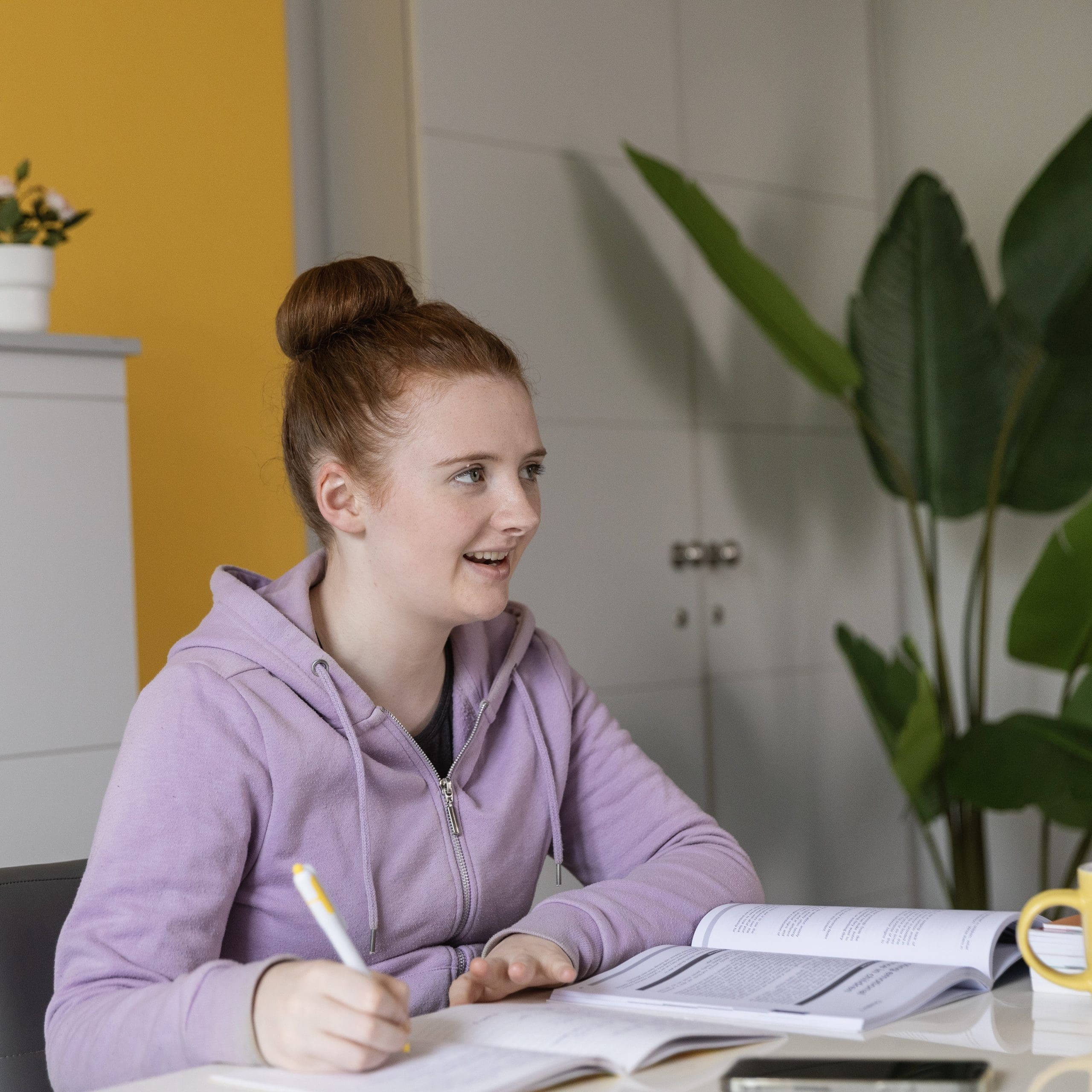 A young woman sits at a desk studying. She is writing in a notebook as she looks to someone out of shot.