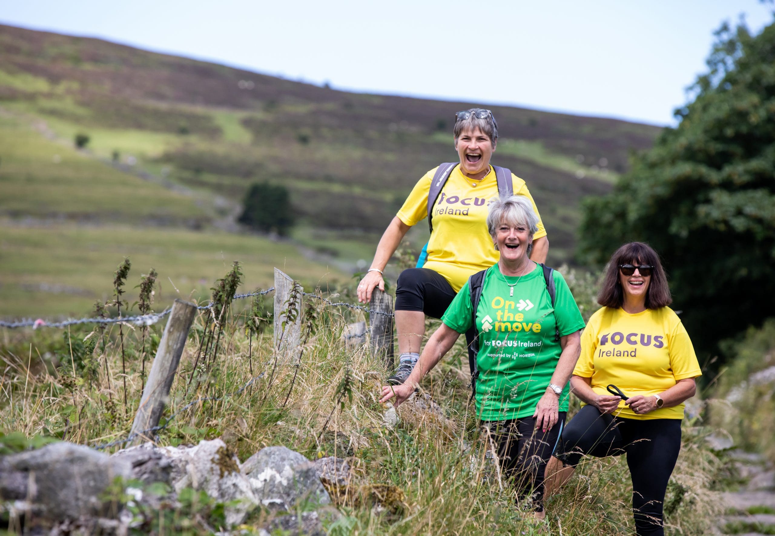 Three hikers smile to camera as they take on Croagh Patrick. Two of them are wearing yellow Focus Ireland t-shirts while the other wears a green on the move t-shirt