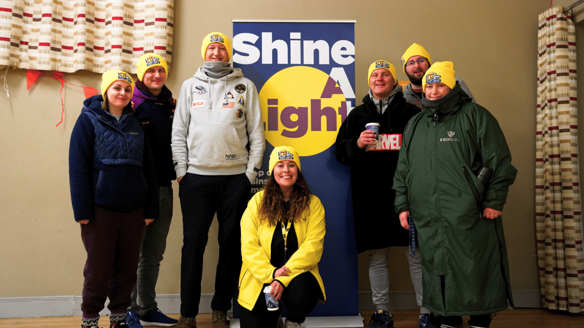 A group of people stand in front of a Shine A Light banner. They are all wearing yellow beanies