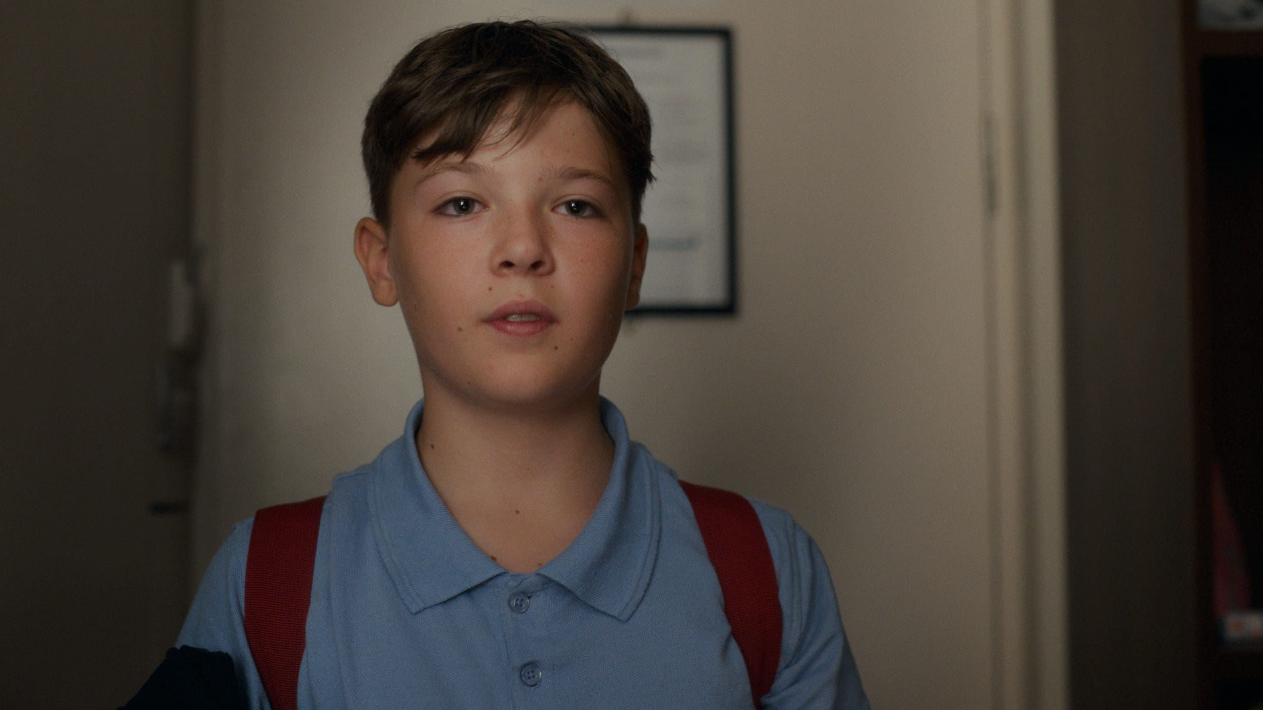 A young boy looks camera. He is wearing a blue polo shirt and wearing a backpack.