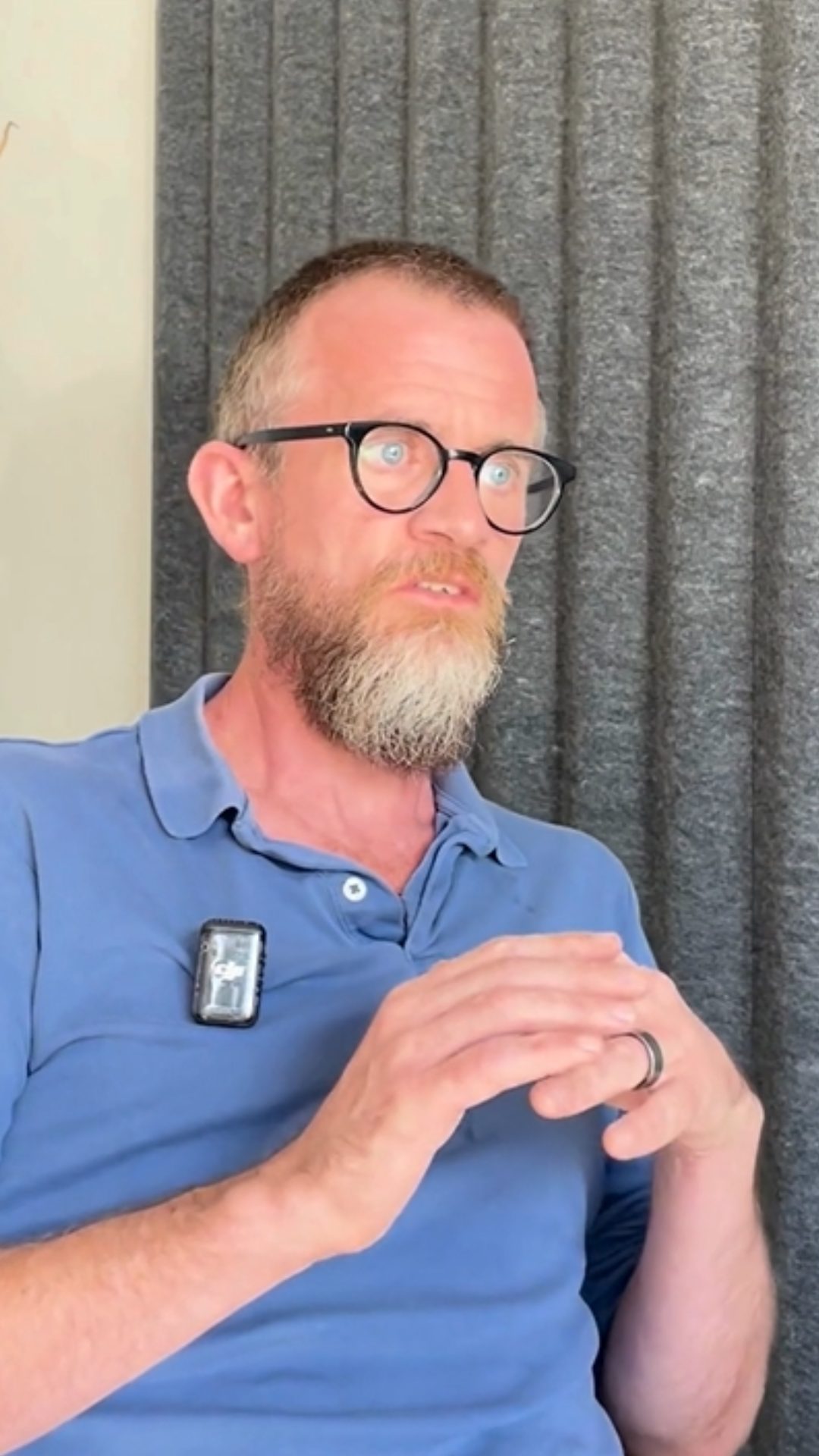 Adrien Quinn speaks to camera. They have a short beard, glasses and are wearing a blue polo top.