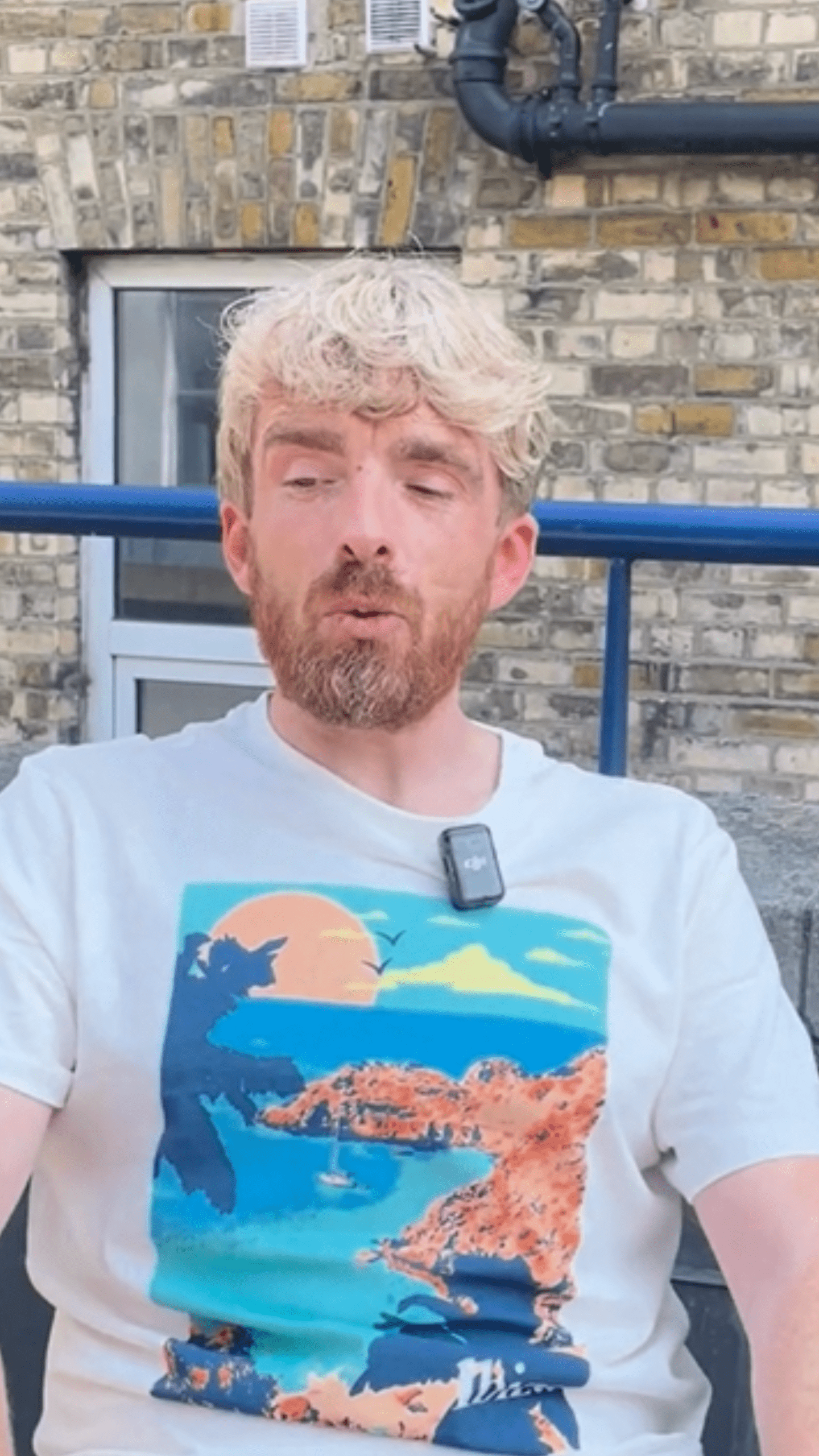 James speaks to camera. They have dyed blonde hair and is wearing a white t-shirt with a print of a beach scene on it.