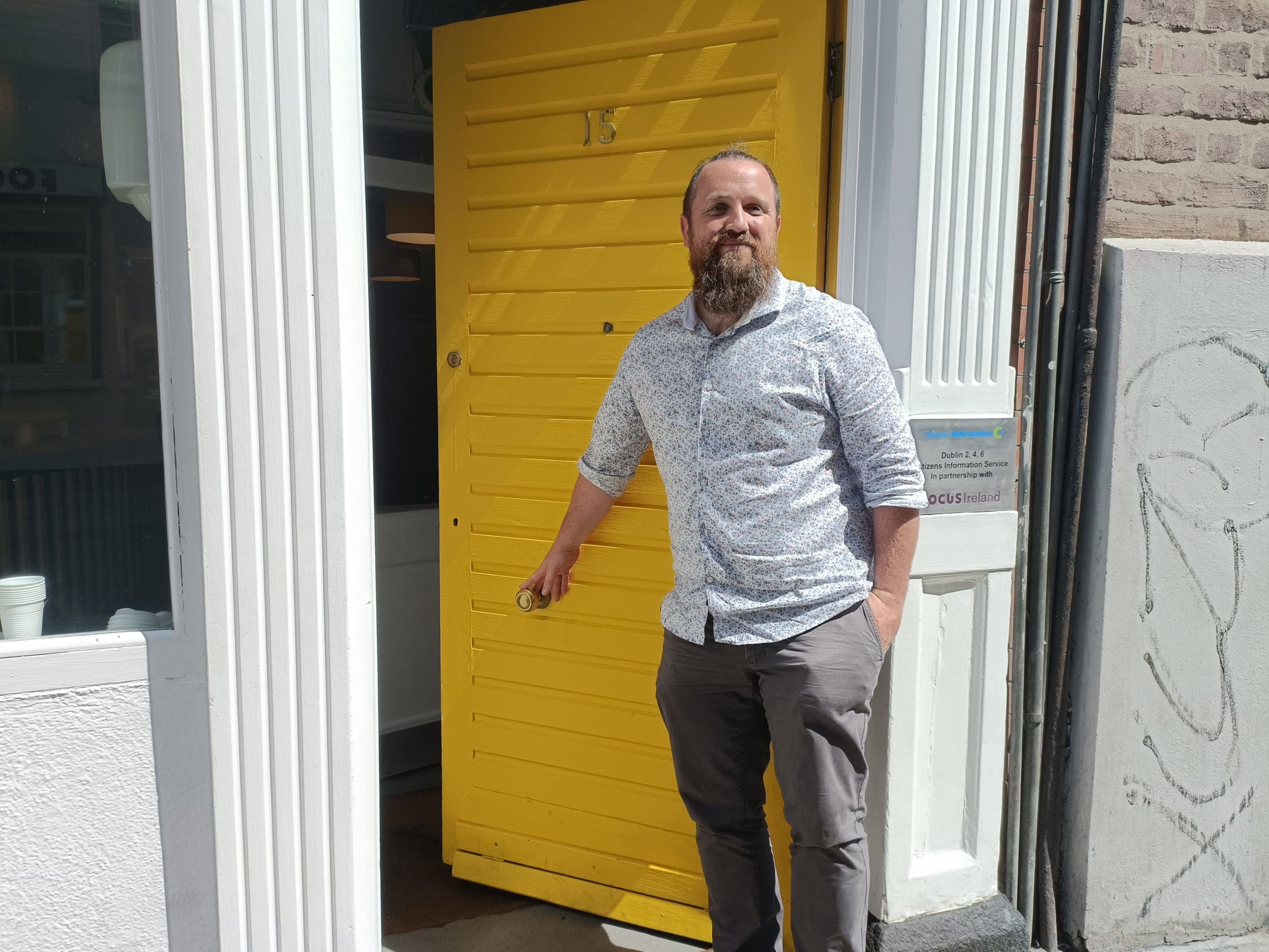 A man in a white shirt stands in front of a bright yellow door
