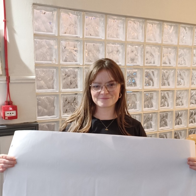 A photo of Rebecca O'Connor. They are holding a large white piece of paper and smiling to camera.