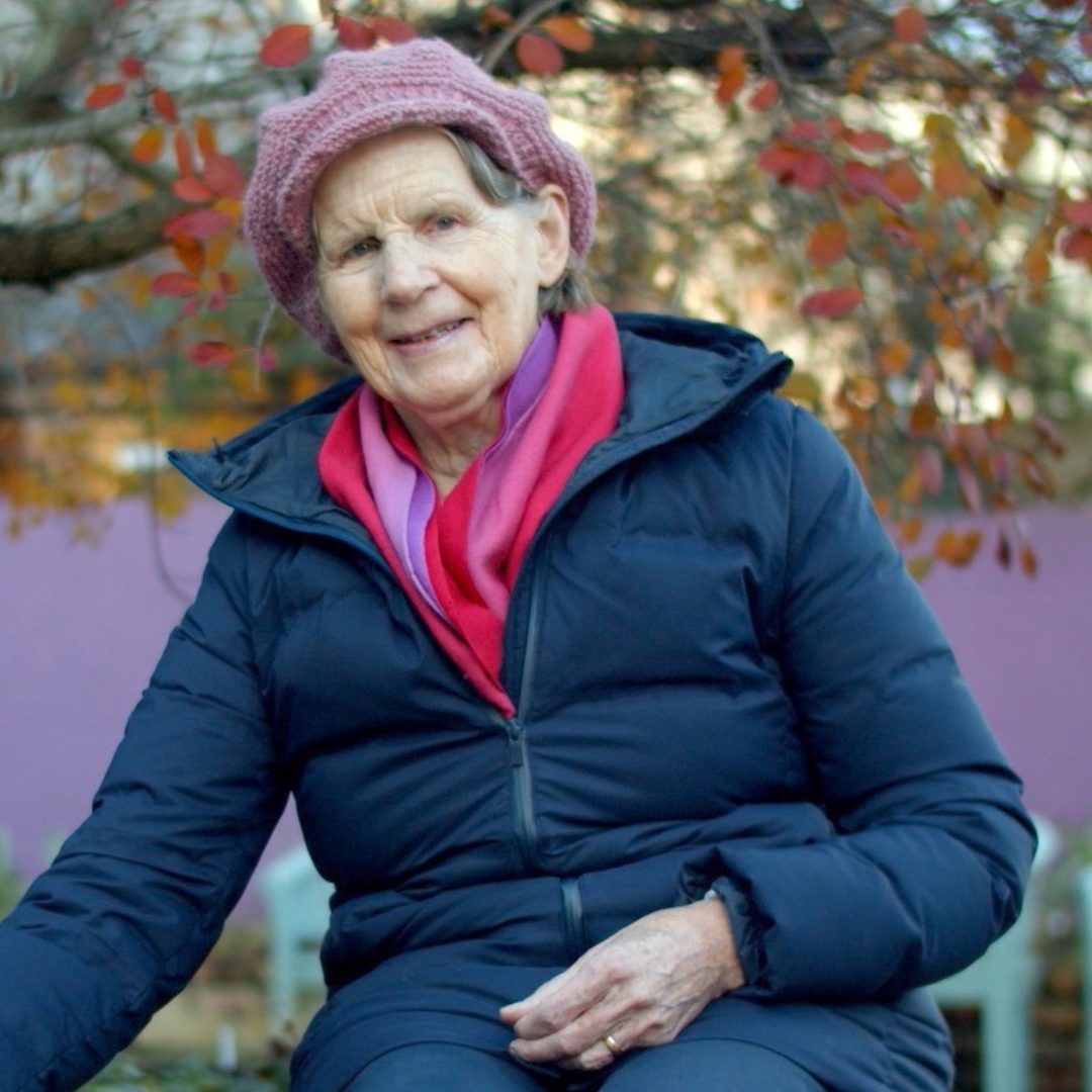 Sister Stan sits in the garden at the sanctuary. They are wearing blue coat and pick wooly hat
